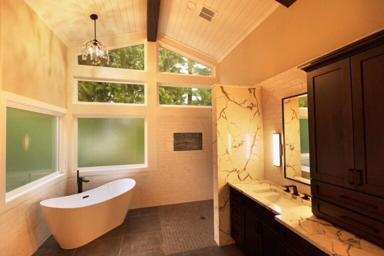 Modern bathroom with high ceilings, large frosted and clear windows, a freestanding white bathtub, a walk-in shower, marble accents, dark wood cabinets, and warm lighting.