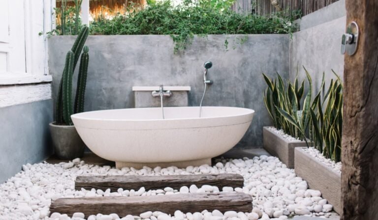 A modern outdoor bathroom with a white freestanding tub on white pebbles, surrounded by green plants, cacti, wooden planks, and gray concrete walls.