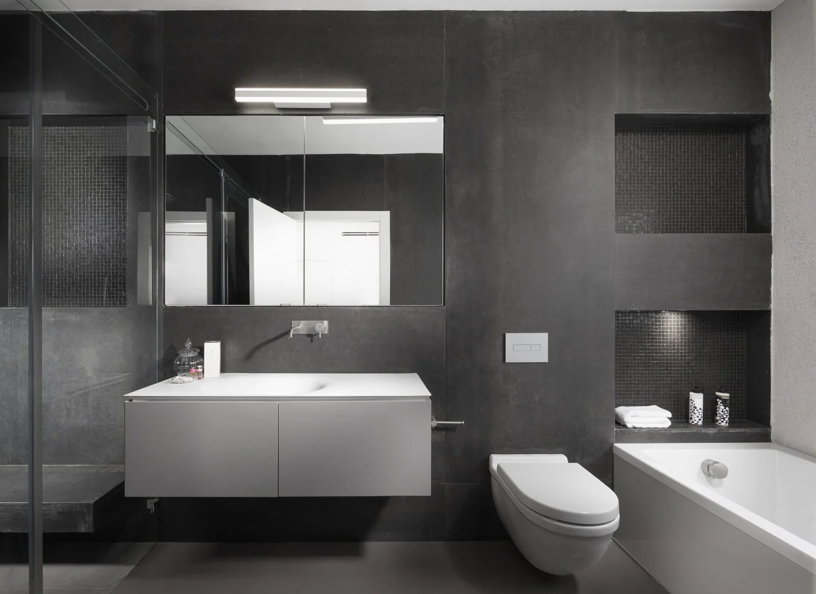 Modern bathroom with dark gray walls, a large mirror above a floating white vanity, and sleek fixtures—an inspiring example of home renovation and remodeling in Seattle. Features include built-in shelving, a wall-hung toilet, and glass shower enclosure.