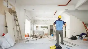 A construction worker wearing a yellow hard hat stands with hands on hips, observing a partially demolished and messy room. Tools, materials, and debris are scattered—raising the question: Do I need an architect for a home renovation in Shoreline?.