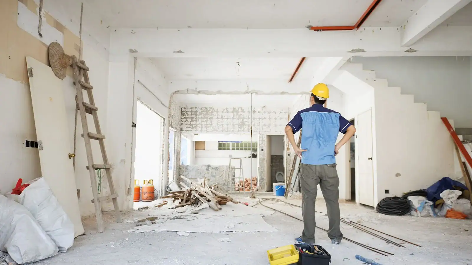 A construction worker wearing a yellow hard hat stands with hands on hips, observing a partially demolished and messy room. Tools, materials, and debris are scattered—raising the question: Do I need an architect for a home renovation in Shoreline?.