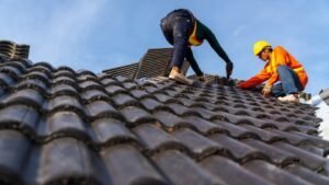 Two men working together on a tiled roof, carefully handling tools and roofing materials. They appear to be repairing or installing clay or concrete tiles, ensuring proper alignment and secure placement for a durable finish.