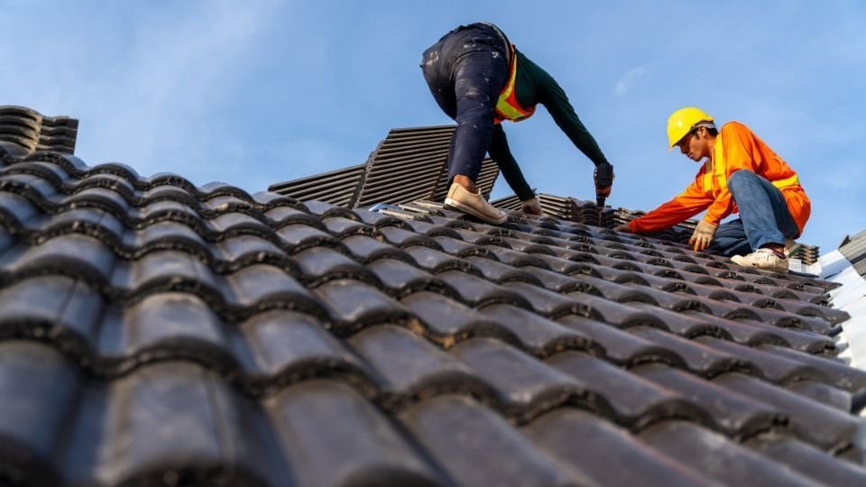Two men working together on a tiled roof, carefully handling tools and roofing materials. They appear to be repairing or installing clay or concrete tiles, ensuring proper alignment and secure placement for a durable finish.