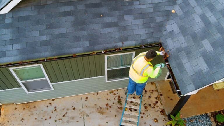 A man is actively working on the roof of a house, equipped with tools for construction or repair. He is positioned carefully on the sloped surface, focusing on roofing tasks such as installation, maintenance, or inspection. The scene highlights skilled labor and home improvement work at height.