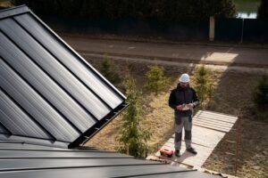 A man is standing on top of a house with a metal roof, possibly inspecting or preparing for maintenance work. The roof’s shiny panels reflect light, emphasizing the durability and modern style of the metal roofing system. His stance suggests focus and awareness of safety while working at height.