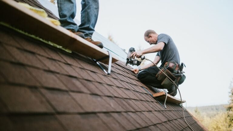 Two men are working on the roof of a house with a ladder leaning against the structure for access. Both are engaged in roofing tasks such as installing or repairing shingles, showcasing teamwork and construction skills. The setup highlights the use of proper tools and equipment in roof maintenance.