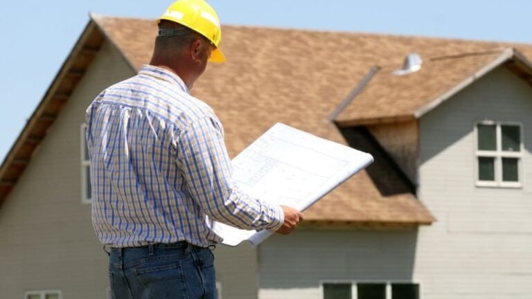 A man standing in front of a house while holding a blueprint. He appears to be reviewing construction or renovation plans, possibly as an architect, contractor, or homeowner. The house is visible in the background, and the blueprint suggests planning or design work in progress.