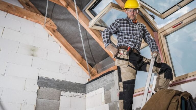 A man wearing a hard hat while standing on a ladder, likely engaged in construction, repair, or maintenance work. He is dressed in work attire, and the ladder is securely positioned against a structure such as a house, roof, or wall. The hard hat emphasizes safety during the task.
