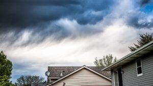 A stormy sky looms over a house with a car parked in front. Dark clouds gather above, creating a dramatic and moody atmosphere. The house stands beneath the unsettled sky, while the car in the driveway adds a sense of everyday life amid the approaching storm.