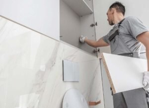 A man standing in front of a toilet, holding a piece of paper, possibly reading instructions or reviewing information related to plumbing or installation. He is wearing casual clothes, and the bathroom features standard fixtures including the toilet and surrounding walls.
