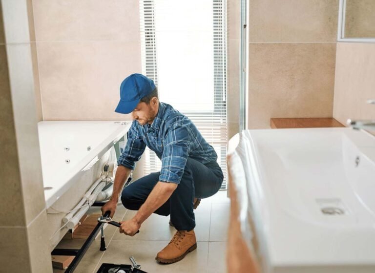 A man repairing a bathroom sink, using tools to adjust or fix plumbing underneath the basin. He is wearing work clothes, and the bathroom features standard fixtures such as a faucet, countertop, and mirror. The scene highlights hands-on maintenance or plumbing work.