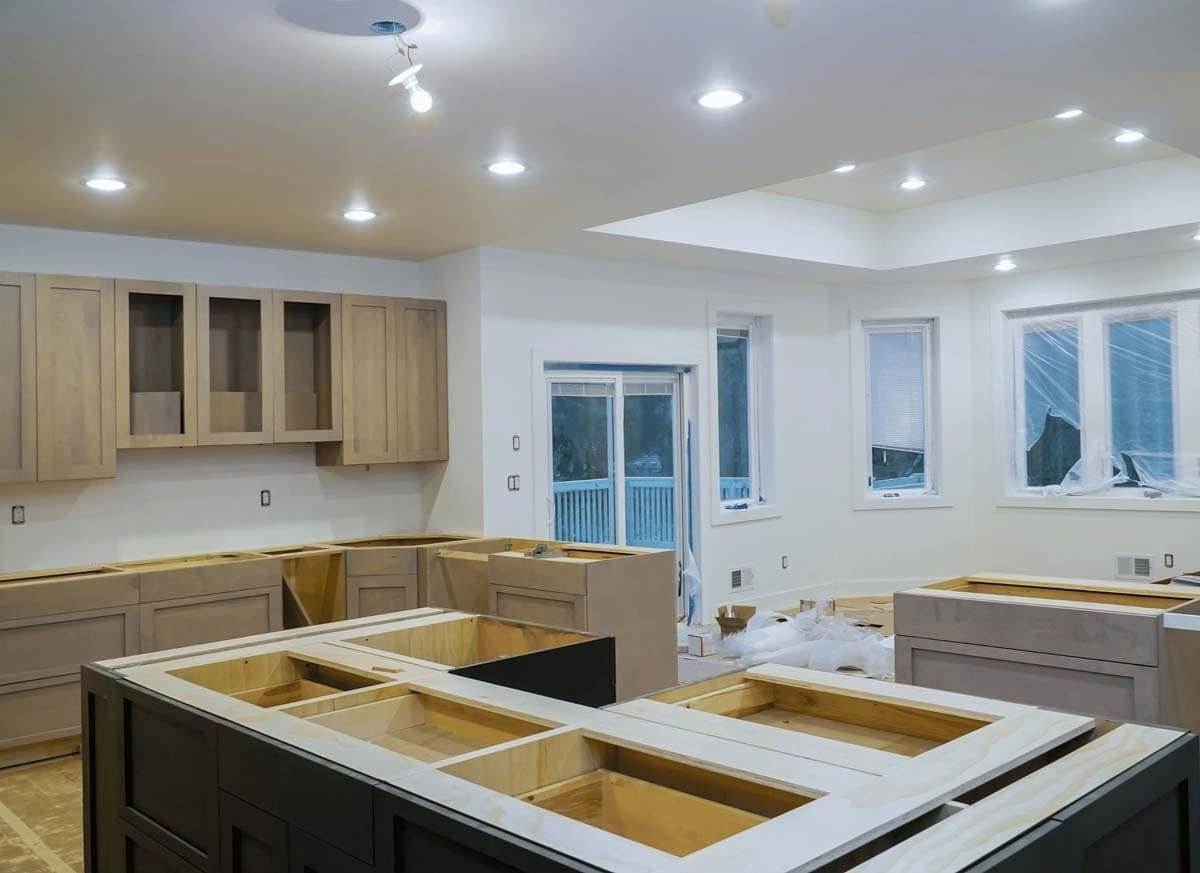 A kitchen under construction with cabinets and countertops being installed. Tools, building materials, and partially assembled cabinetry are visible, highlighting the ongoing renovation or new kitchen build. The space shows unfinished surfaces and an open layout, emphasizing the work-in-progress.