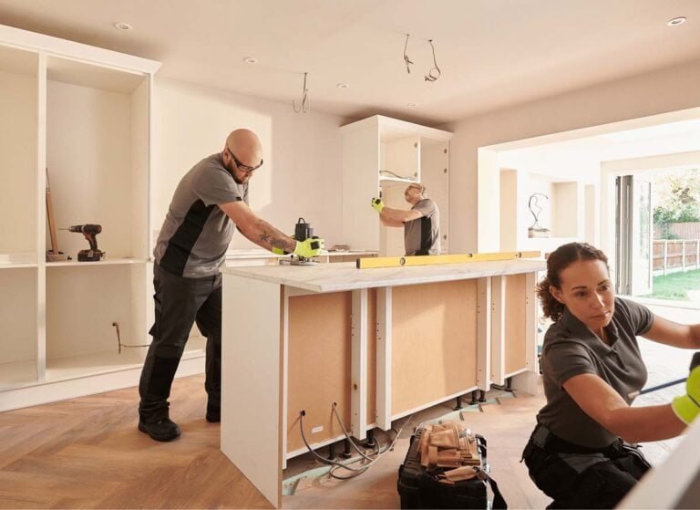Two people working together on a home kitchen renovation. One person may be installing cabinets while the other works on countertops or appliances. Tools and construction materials are visible around the space, and the kitchen is partially built, highlighting an active remodeling project.