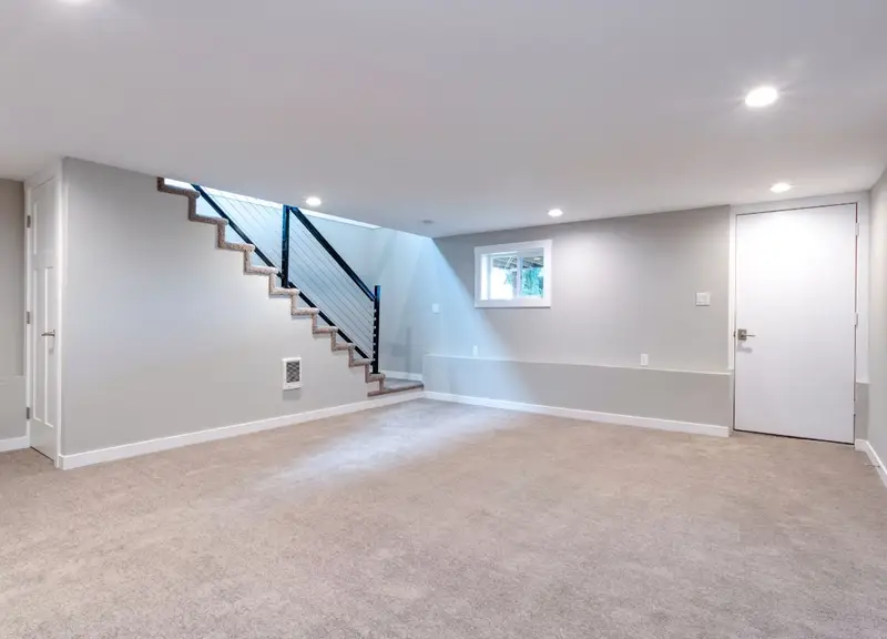 A clean, well-lit basement featuring a set of stairs leading down to the space and a closed door at the far end. The basement floor and walls are tidy, with minimal clutter, giving the area an organized and spacious appearance. The lighting highlights the stairs, door, and surrounding floor space.