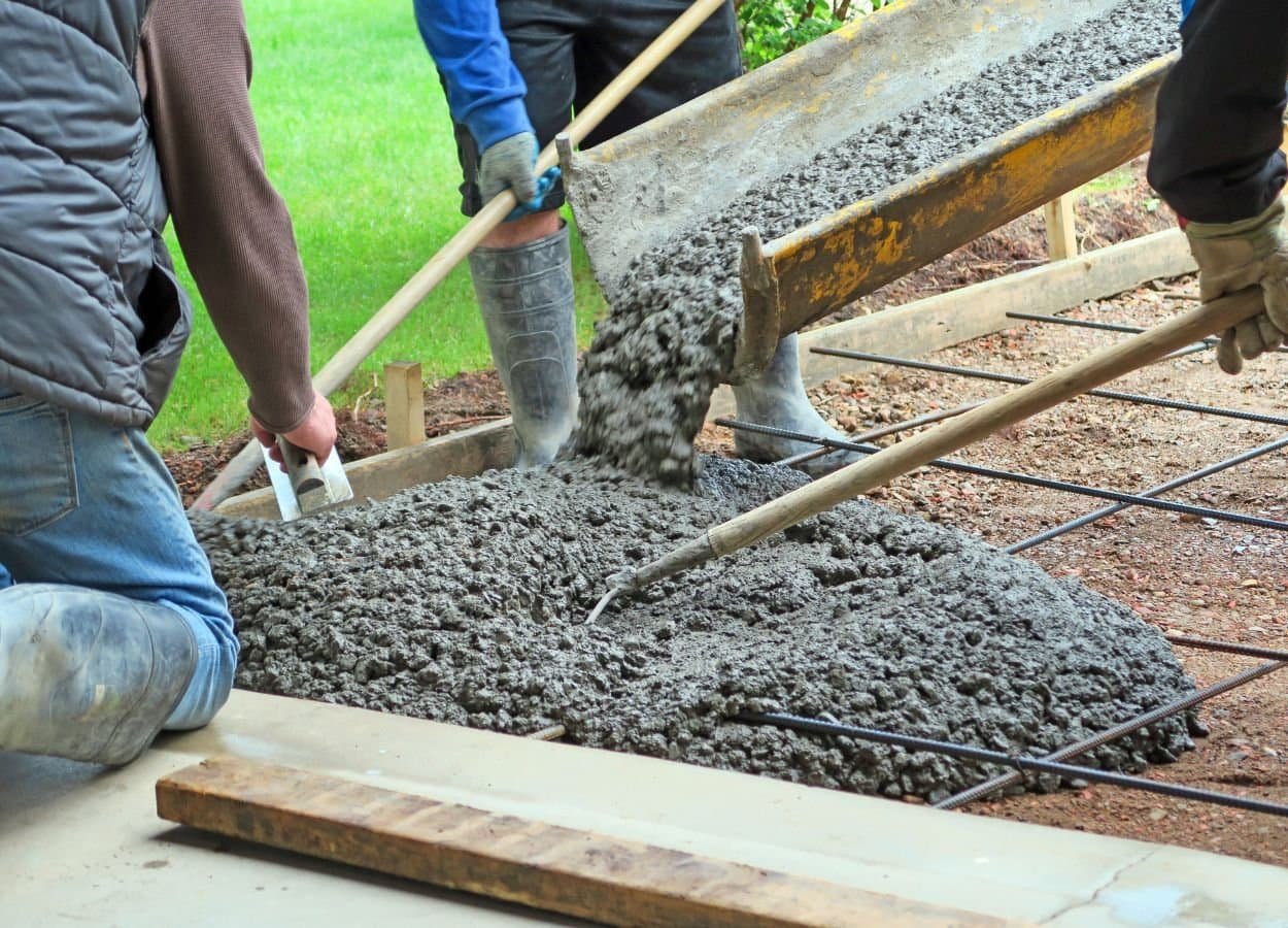 Concrete being poured in a backyard area, spreading over a prepared surface to create a patio, walkway, or slab. Construction tools and equipment are visible around the work site, and the surrounding yard includes grass, plants, or fencing. The scene captures the process of backyard renovation or construction.