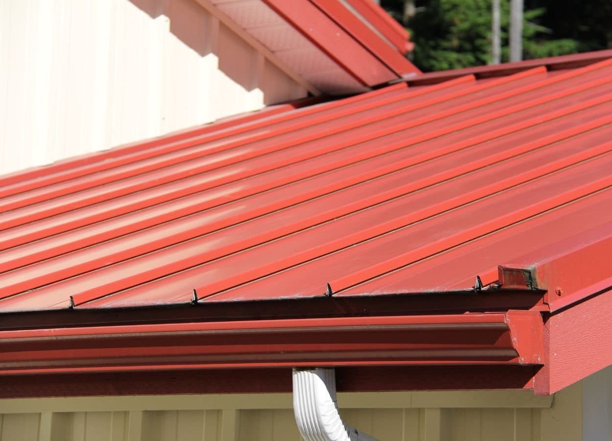 A house roof made of red metal panels with a crisp white trim along the edges. The metal roof has a modern, shiny finish, contrasting sharply with the white trim. The house exterior is partially visible, highlighting the clean architectural lines and vibrant color combination.