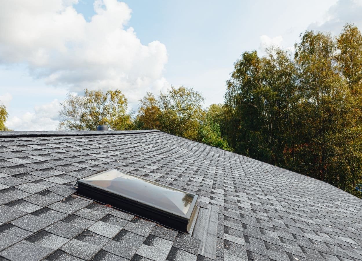 A roof featuring a skylight that allows natural light to enter the space below. The roof is covered with neatly arranged shingles or panels, and the skylight is framed in a clean, modern style. Sunlight reflects subtly off the skylight glass, enhancing the aesthetic and functional appeal of the roof.