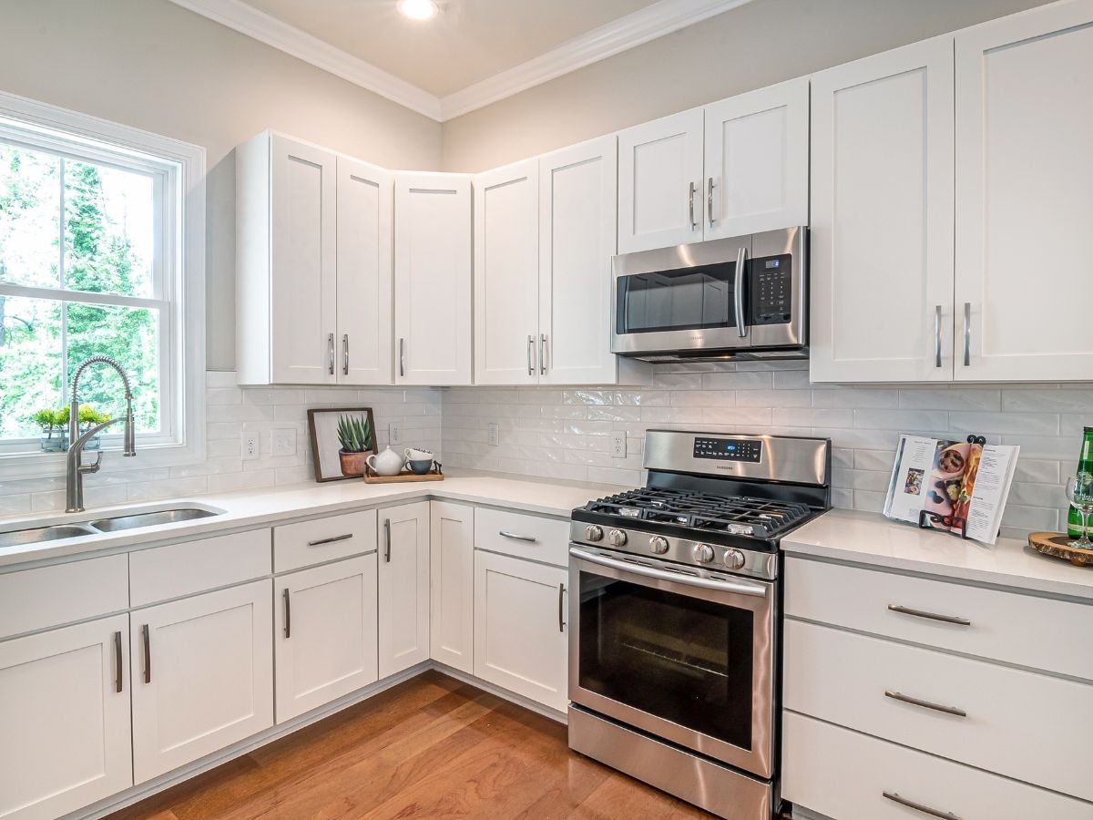 A modern kitchen featuring white cabinetry paired with stainless steel appliances, including a refrigerator, stove, and oven. The space has clean lines, a sleek countertop, and possibly an island or open shelving, creating a bright and functional environment. Lighting highlights the reflective surfaces of the stainless steel and the crisp white cabinets, emphasizing contemporary design.