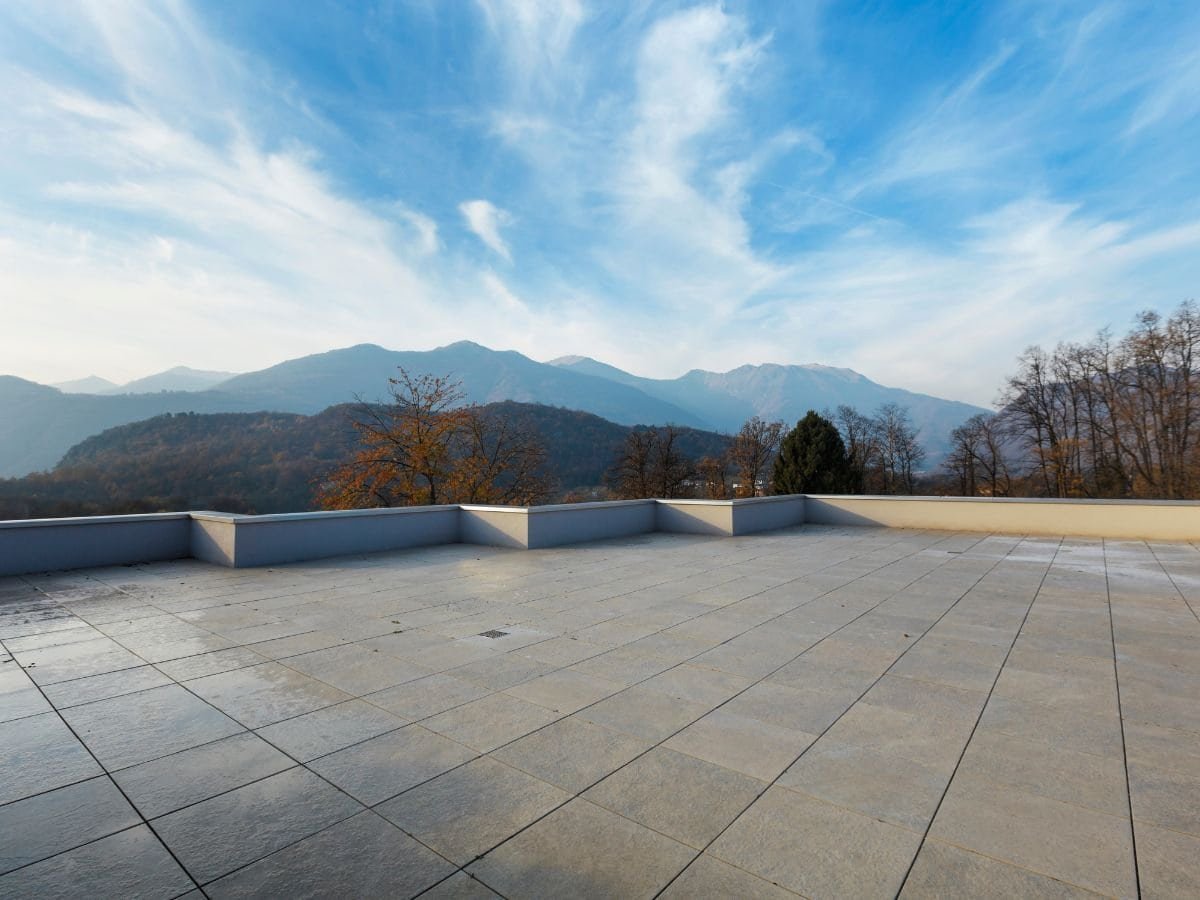 A scenic view of distant mountains as seen from a rooftop. The roof in the foreground features flat or sloped surfaces, while the mountains in the background rise majestically under a clear or partly cloudy sky. The scene captures a blend of urban or residential architecture with natural landscapes, emphasizing perspective and depth.