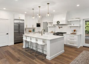 A bright, modern kitchen featuring all-white cabinets and countertops, centered around a spacious island. The kitchen includes a sink, stove, and appliances, with clean lines and minimalist fixtures. Natural or artificial lighting highlights the glossy surfaces and open layout, creating an airy, functional, and elegant environment.