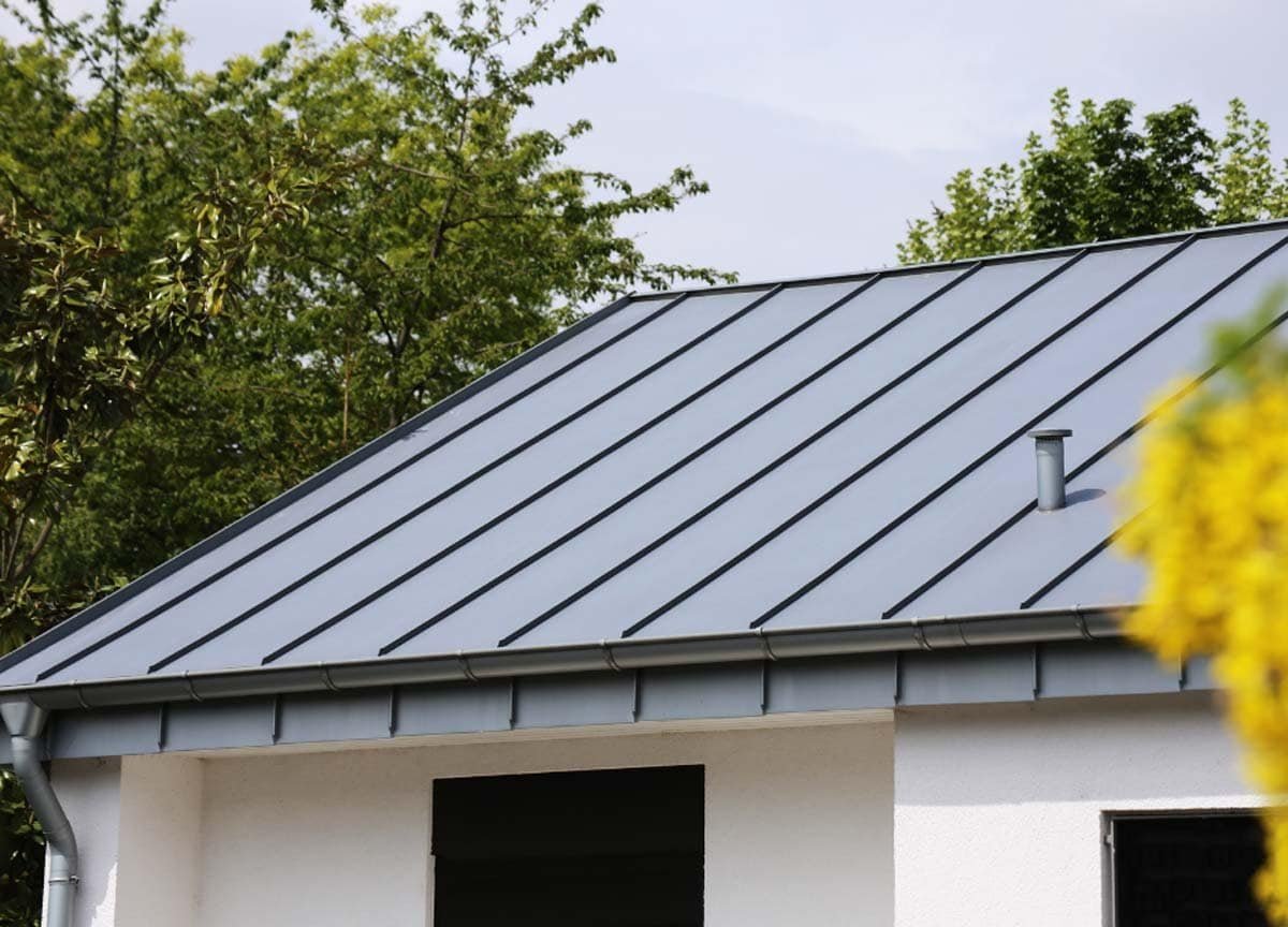 A house with a shiny metal roof and a visible window on the front side. The metal panels are neatly aligned, giving the roof a modern and durable look. The window has a clean frame and reflects the surrounding environment, complementing the house’s contemporary exterior.