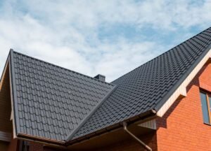 A modern house featuring a sleek metal roof, reflecting sunlight with a subtle shine. The house has large windows and a well-maintained exterior, surrounded by a green lawn and small shrubs. The metal roofing panels are arranged neatly, giving the house a contemporary and durable appearance.