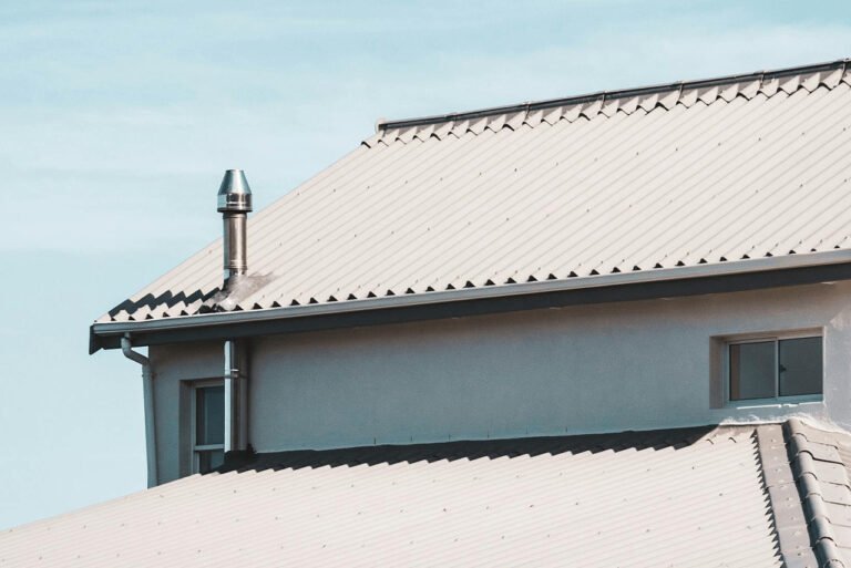 A house roof featuring a sleek white metal surface with a prominent chimney. The roof has clean lines and a modern appearance, reflecting sunlight and adding a bright, polished look to the building. The chimney rises from the roof, complementing the structure’s overall design.