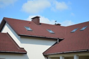 A charming house featuring a vibrant red roof and multiple windows with white frames. The exterior walls are light-colored, and the house is surrounded by greenery, creating a warm and inviting appearance. The red roof adds a striking contrast to the overall design.