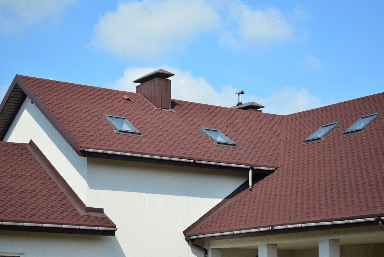 A charming house featuring a vibrant red roof and multiple windows with white frames. The exterior walls are light-colored, and the house is surrounded by greenery, creating a warm and inviting appearance. The red roof adds a striking contrast to the overall design.