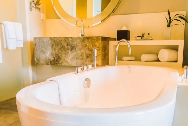 A bathroom featuring a bathtub positioned in front of a large mirror that spans part of the wall. The space may include a sink and other fixtures, with tiled or painted walls and flooring. The mirror reflects light, making the room appear spacious and bright.