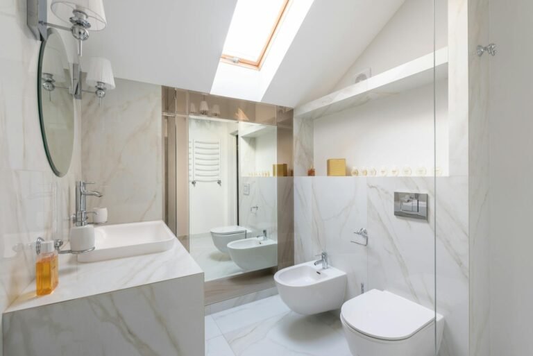 "A luxurious modern bathroom featuring white marble walls and floor, creating a clean and elegant atmosphere. The space includes a contemporary sink with a sleek countertop, a wall-mounted toilet, and a glass-enclosed shower. Subtle recessed lighting enhances the natural shine of the marble, highlighting the sophisticated and minimalist design.