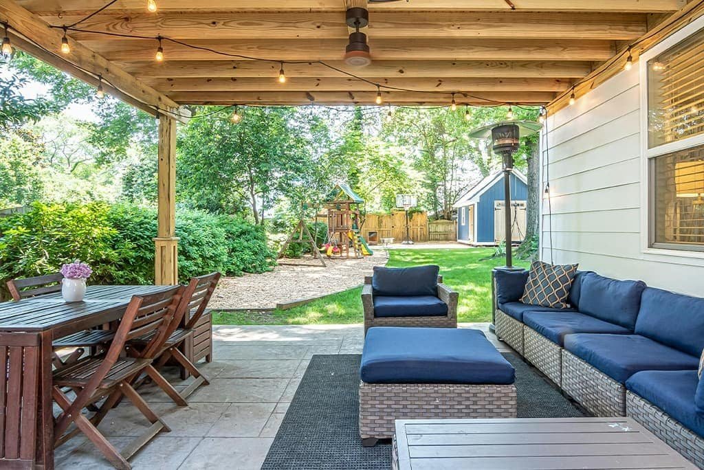 "A covered outdoor patio featuring blue cushioned couches arranged around a table. The area is shaded by a roof or pergola, creating a comfortable seating space for relaxation or social gatherings. Sunlight may filter in, highlighting the furniture and the inviting ambiance of the patio.