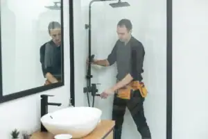 A man wearing a black shirt stands in front of a bathroom sink with a mirror above it. The sink area features white fixtures and a clean countertop, while the reflection in the mirror captures part of the bathroom interior. The man appears to be preparing for or finishing a daily routine.