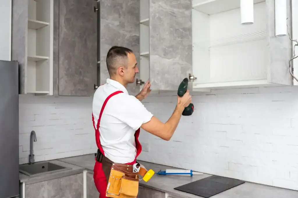 A man wearing a work apron is fixing cabinets in a modern kitchen. He is adjusting the cabinet doors with a screwdriver, ensuring proper alignment and fit. The bright kitchen setting features clean countertops and neatly arranged fixtures, highlighting a home improvement project in progress.
