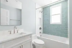 A modern bathroom featuring blue ceramic tile walls and floor, paired with sleek white fixtures. A white sink, toilet, and bathtub stand out against the bold tile, while chrome accents and bright lighting enhance the clean, fresh look of the space.
