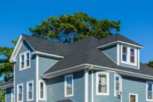 A two-story blue house with white trim and dark gray shingles, partially surrounded by green trees, under a clear blue sky.
