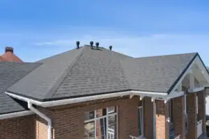A close-up view of a house with a gray shingle roof, brick walls, white trim, and multiple black roof vents against a clear blue sky.