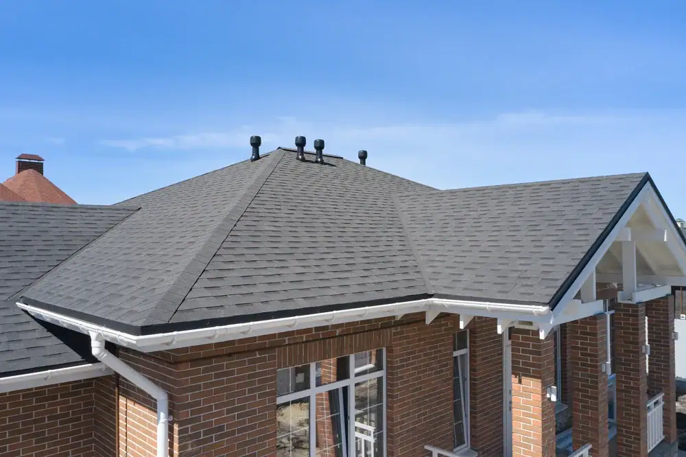 A close-up view of a house with a gray shingle roof, brick walls, white trim, and multiple black roof vents against a clear blue sky.
