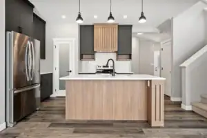 Modern kitchen with a central island featuring light wood tones, black cabinetry, stainless steel appliances, pendant lights, and a white backsplash with geometric patterns. Stairs are visible on the right.