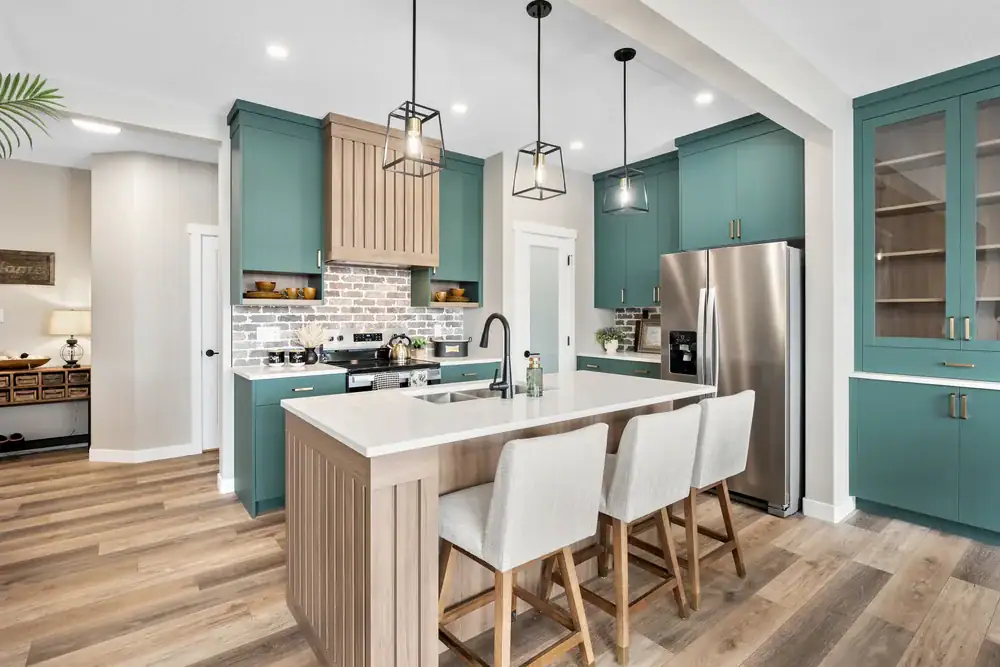 Modern kitchen with teal cabinets, stainless steel appliances, wood accents, and a large island featuring a white countertop and three light-colored upholstered chairs. Pendant lights hang above the island.