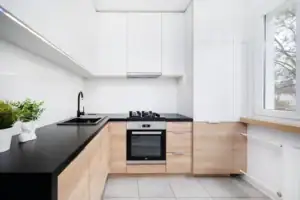 Modern kitchen with white upper cabinets, light wood lower cabinets, black countertops, a built-in oven, gas cooktop, black sink with faucet, potted plants, and a large window letting in natural light.