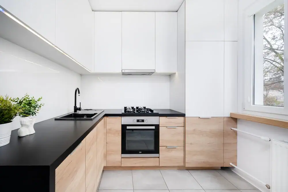 Modern kitchen with white upper cabinets, light wood lower cabinets, black countertops, a built-in oven, gas cooktop, black sink with faucet, potted plants, and a large window letting in natural light.
