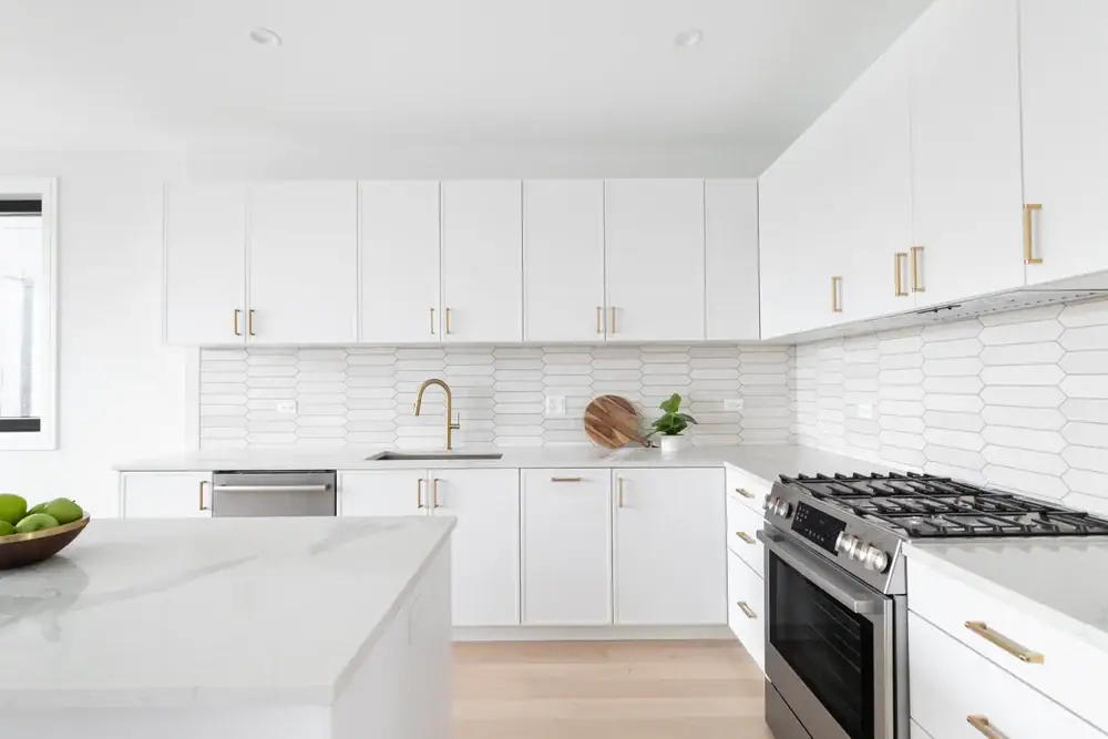 A modern kitchen with white cabinets, gold handles, a gas stove, a dishwasher, a marble countertop, a tile backsplash, a brass faucet, and a bowl of green apples on the island.