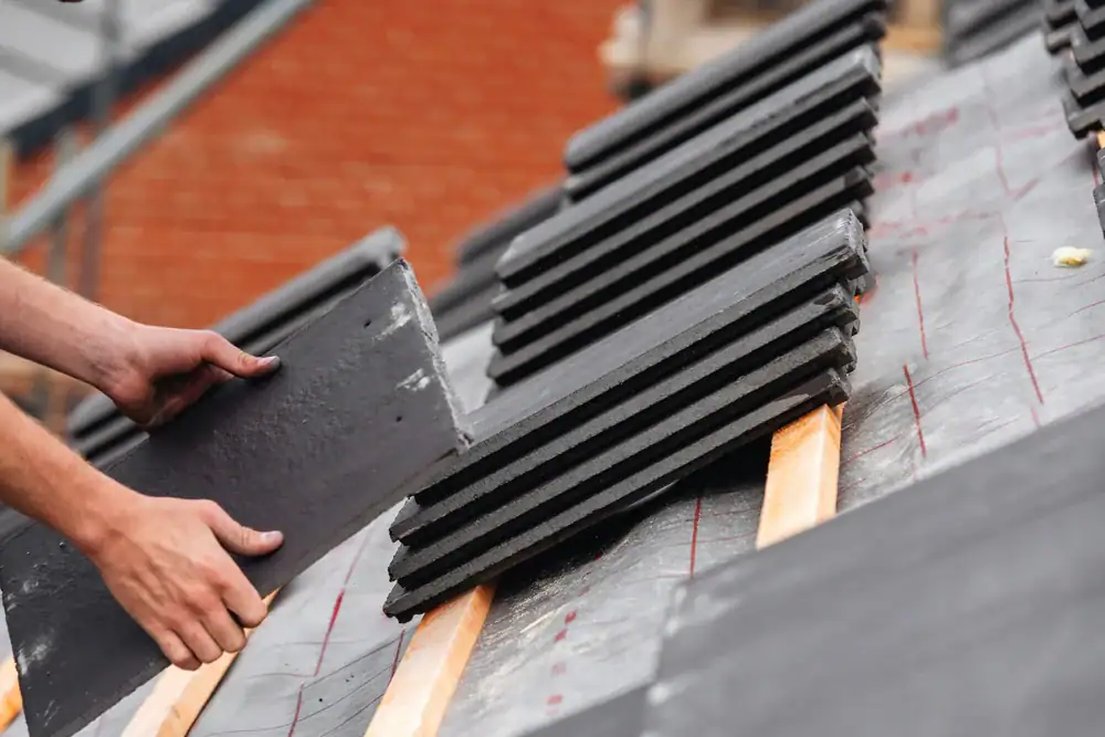 Close-up of a person installing dark roofing tiles on a roof. Several tiles are stacked nearby, and wooden battens are visible underneath. A brick wall is blurred in the background.
