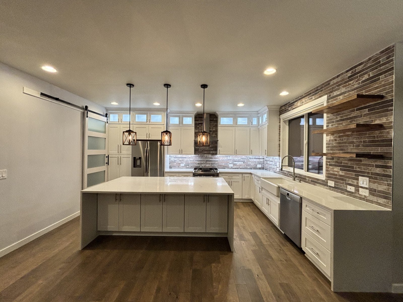 Modern kitchen with white cabinets, large island, stainless steel appliances, pendant lights, wood floors, a sliding frosted glass door, and a brick accent wall with floating shelves—perfect for your Kitchen Renovation Seattle WA.