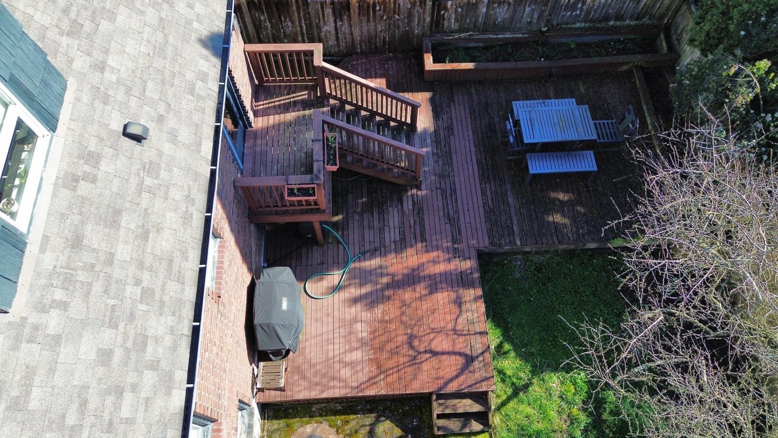 Aerial view of a wooden backyard deck with stairs, patio furniture, and a grill, next to green grass.