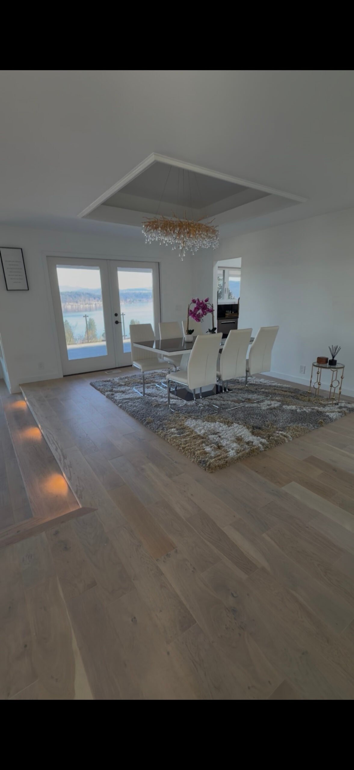 Modern dining room with white chairs, a large rug, wood floors, chandelier, and glass doors to water views.