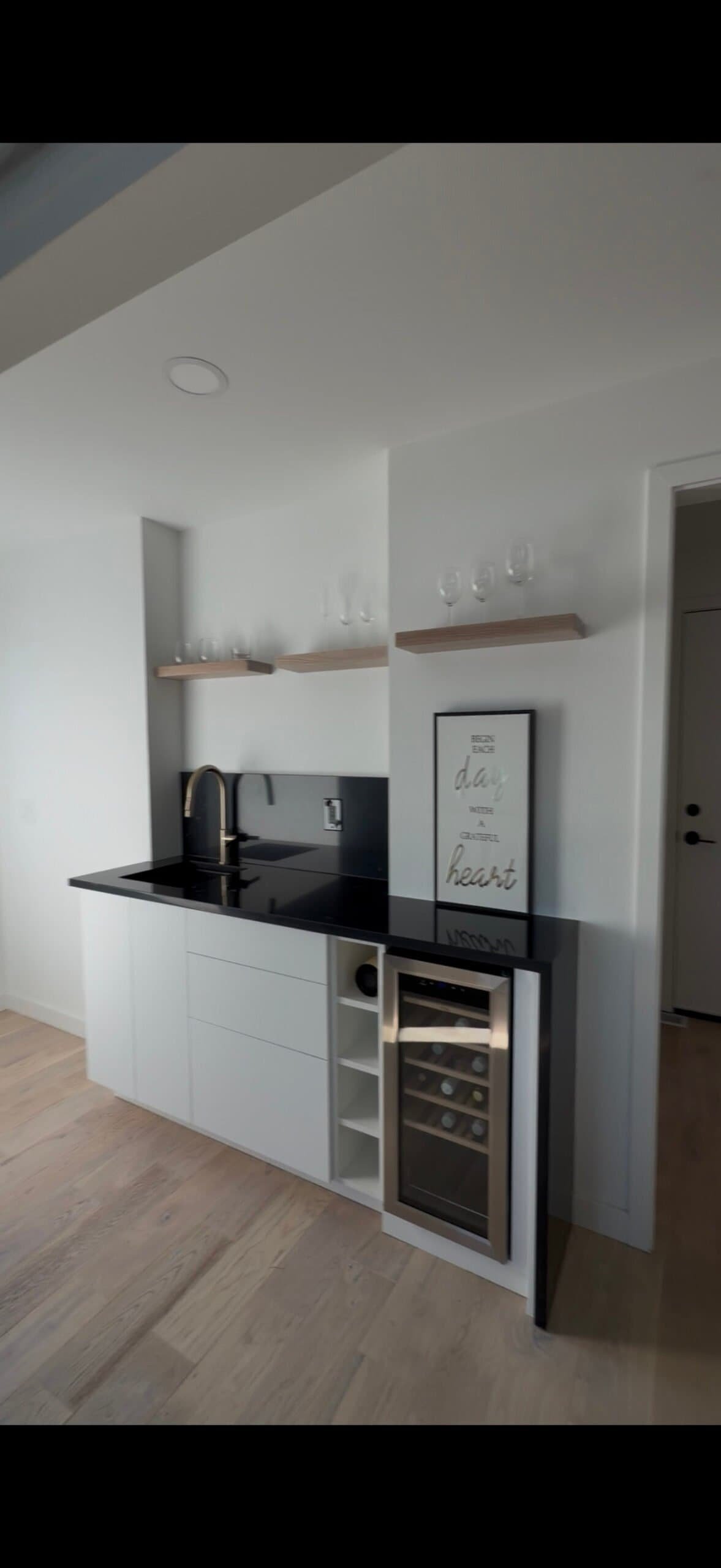 Modern kitchen nook featuring a black countertop, open shelves, wine fridge, and light wood flooring.