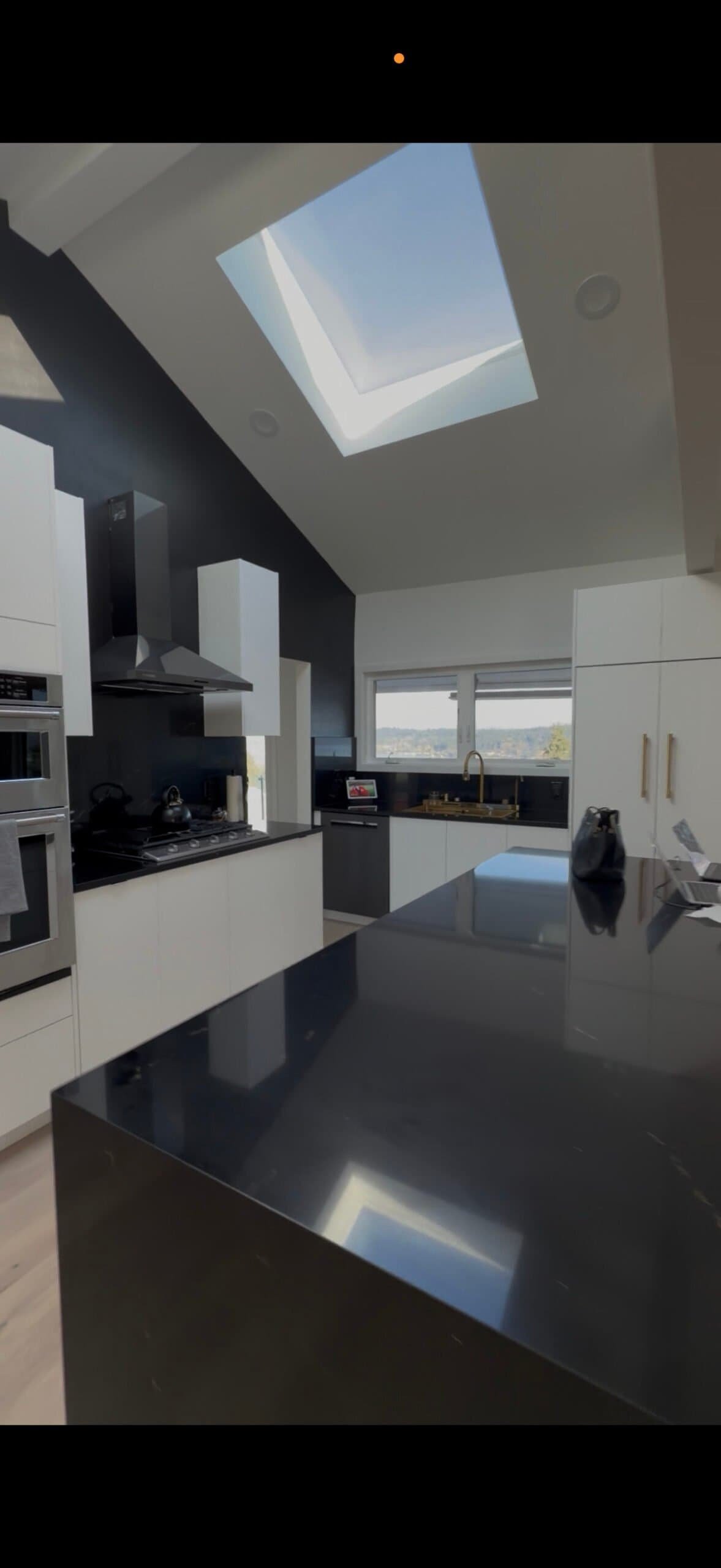Modern kitchen with white cabinets, black countertops, island beneath skylight, and large windows.