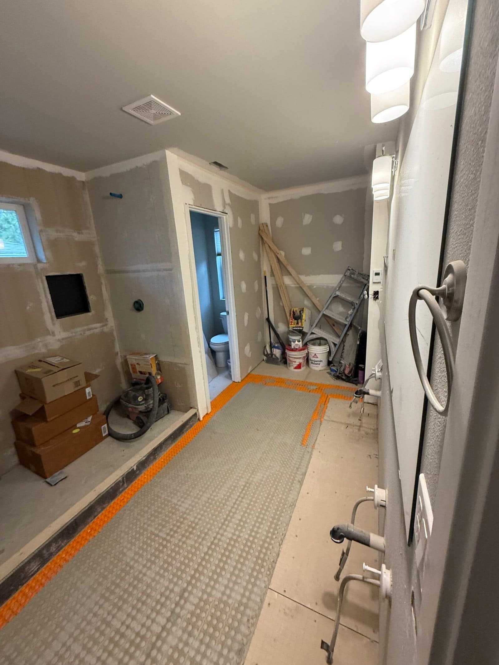 Bathroom under renovation with exposed drywall, tools, boxes, ladder, and unfinished floor and walls.